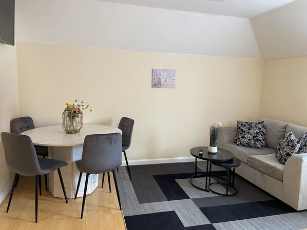 A modern living room with a round dining table, grey upholstered chairs, a grey sofa with patterned cushions, a small side table, and a black and grey rug. - Modern 2BR House Sleeps 6 Parking, Wi-Fi & Self Check-In Near Bluewater & London (Greenhithe)