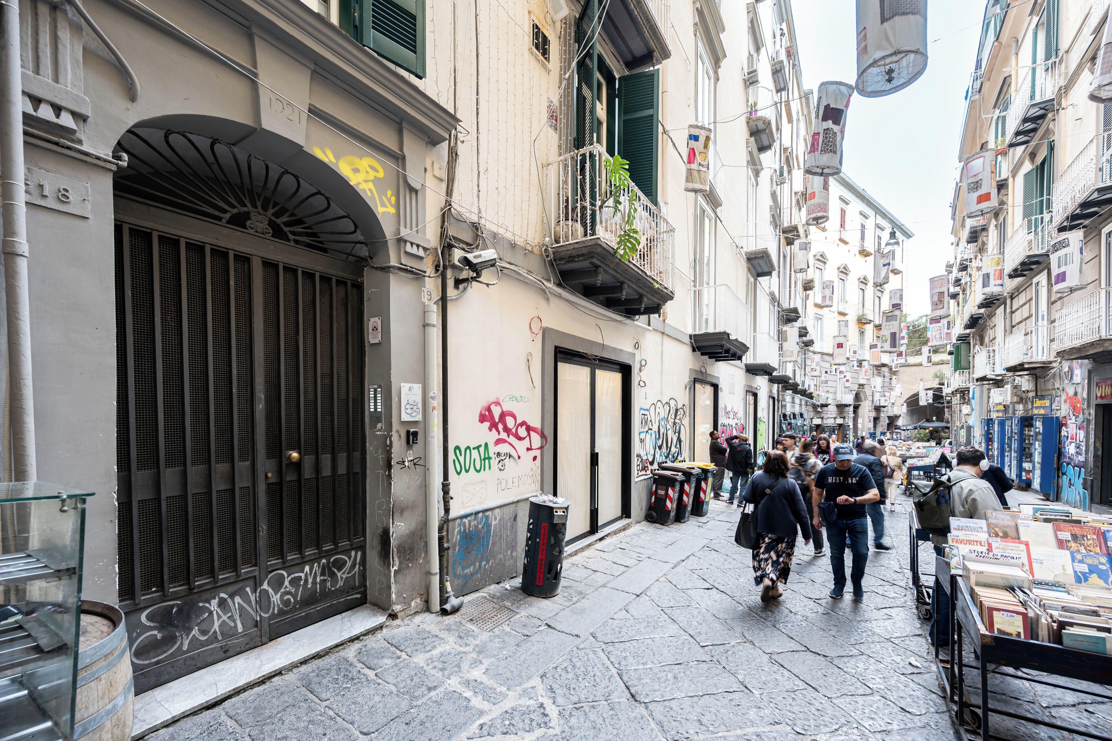 A narrow street with buildings, a closed shop with graffiti, and a street vendor with books.