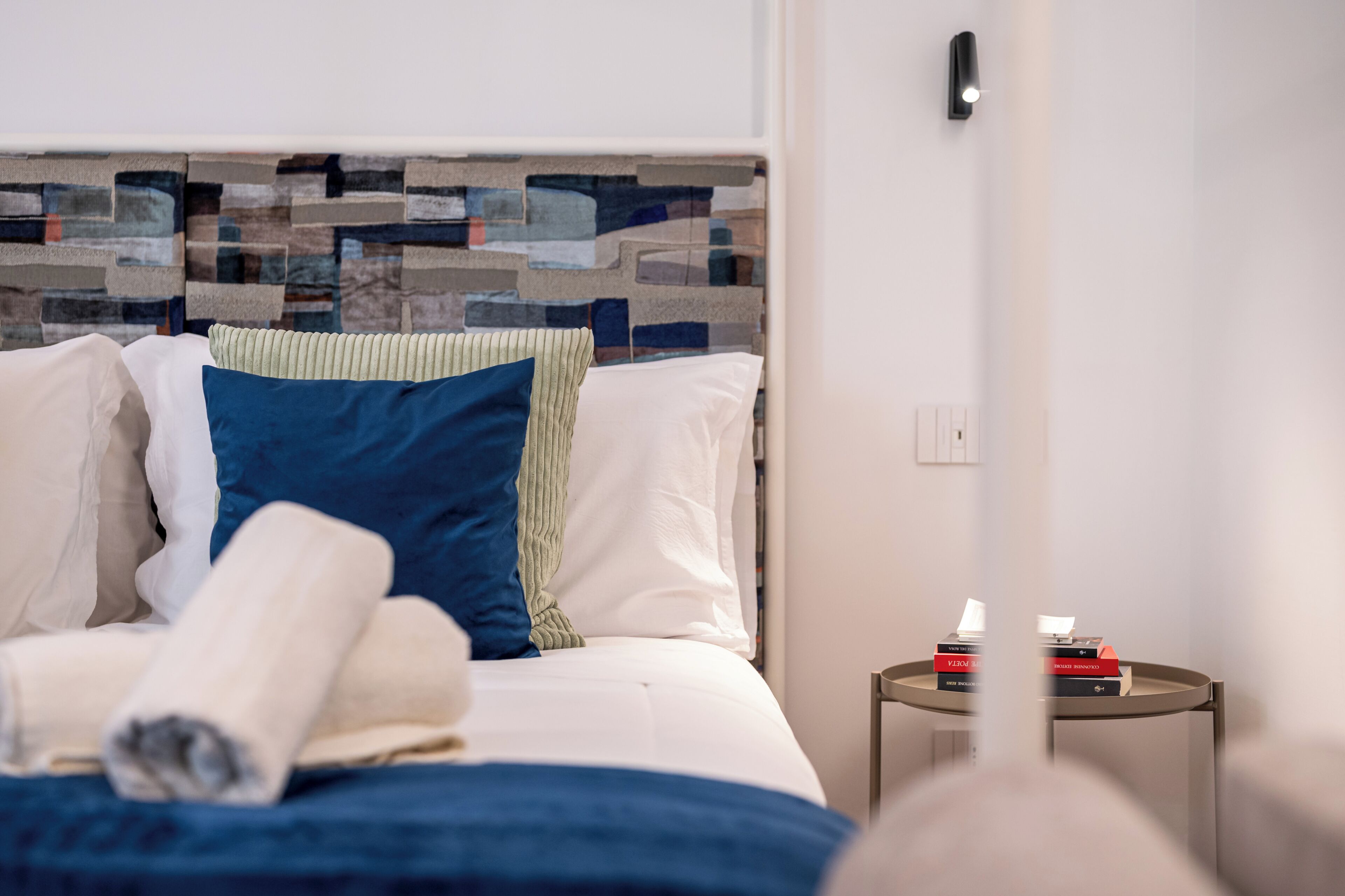 A neatly made bed with pillows and a rolled towel, a small side table with books, and a modern wall with a textured design.