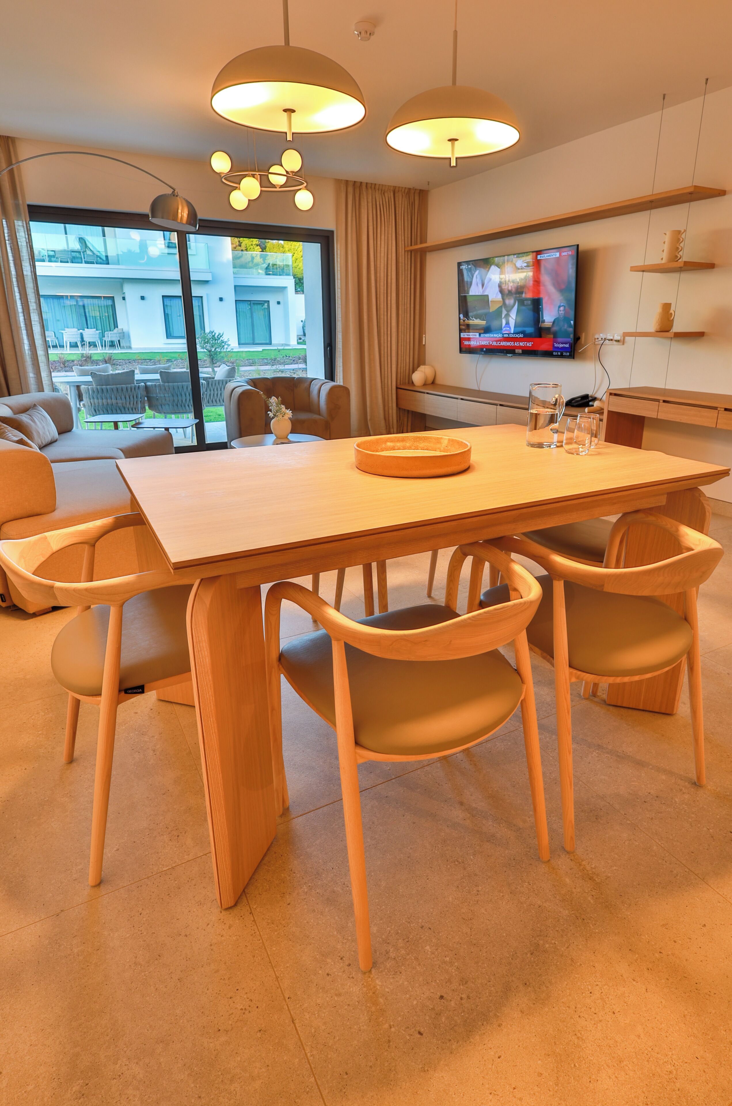 A modern dining area with a wooden table and chairs, a television mounted on the wall, and a view of the outdoors.
