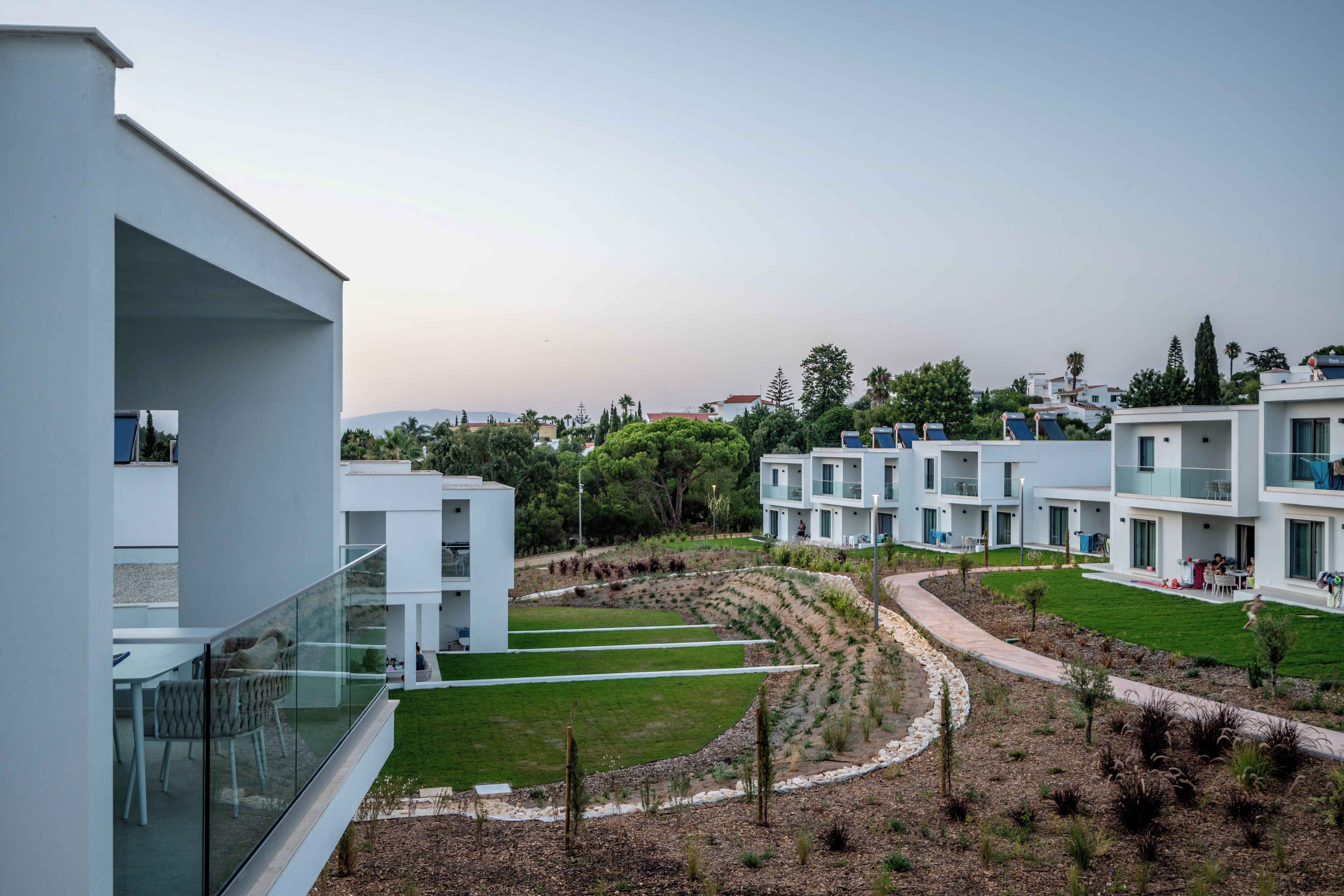 A modern residential complex with multiple white buildings, a landscaped garden, and a clear sky.