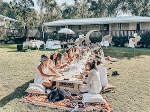 A group of people seated at an outdoor dining table with a white tablecloth, surrounded by a grassy area with a building in the background. - Hunter Moon - A stunning Country Estate (Broke)
