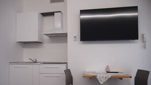 Een compacte keuken met een ingebouwde oven, een magnetron en een spoelbak. Er is een kleine eettafel met twee stoelen en één vaas. Een flatscreen televisie is aan de muur boven de tafel bevestigd.