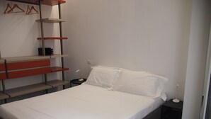 Een net opgemaakt bed met witte lakens, een nachtkastje met een lamp, en een wandplank met een koffieapparaat.