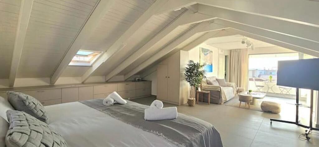 Quarto de hotel com uma cama, área de estar com mesa e cadeiras, televisão e varanda com vista.