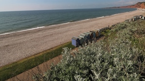 A beach with a row of small, uniform buildings, a pebbled beach, and a clear view of the ocean. - The Cottage (Budleigh Salterton)