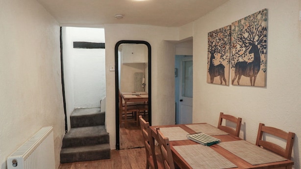 A dining area with a wooden table and chairs, a staircase, and a hallway leading to another room. - The Cottage (Budleigh Salterton)