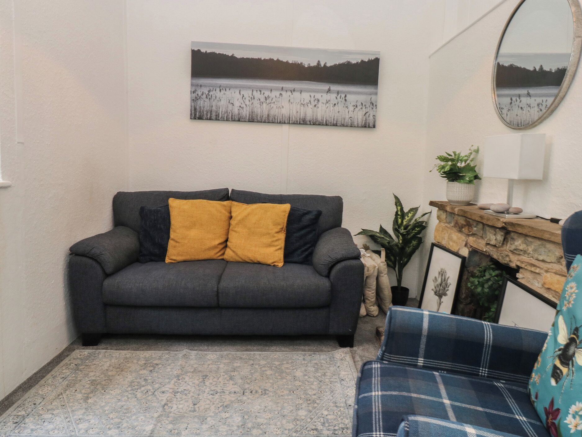 Una sala de estar con un sofá gris, un sillón azul, un cojín amarillo, un cuadro en la pared y una pequeña planta en maceta.