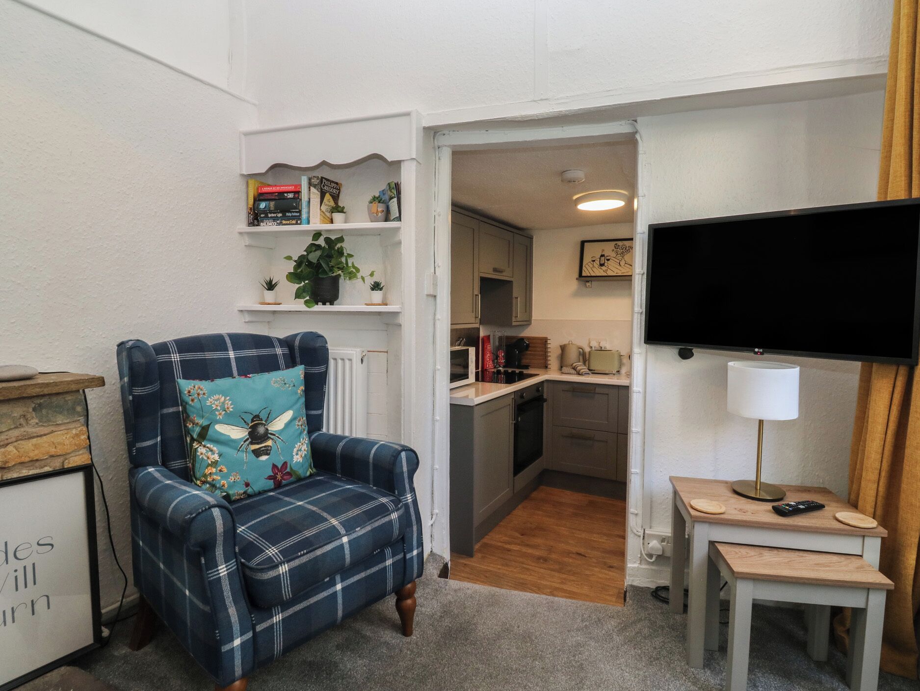 Una sala de estar con un sillón azul a cuadros, un televisor de pantalla plana, una mesita de noche con lámpara y una cocina al fondo.
