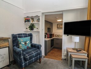 A living room with a blue plaid armchair, a flat-screen TV, a small table with a lamp, and a kitchen area in the background. - The Cottage (Budleigh Salterton)