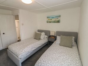 A bedroom with two single beds, a nightstand with a lamp, a painting on the wall, and a door leading to another room. - The Cottage (Budleigh Salterton)