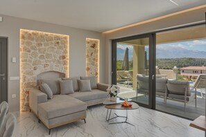 Ein modernes Wohnzimmer mit einer Wand aus Stein, einem Ecksofa, einer Glastür, die auf einen Balkon mit Sitzgelegenheit und Bergblick führt.