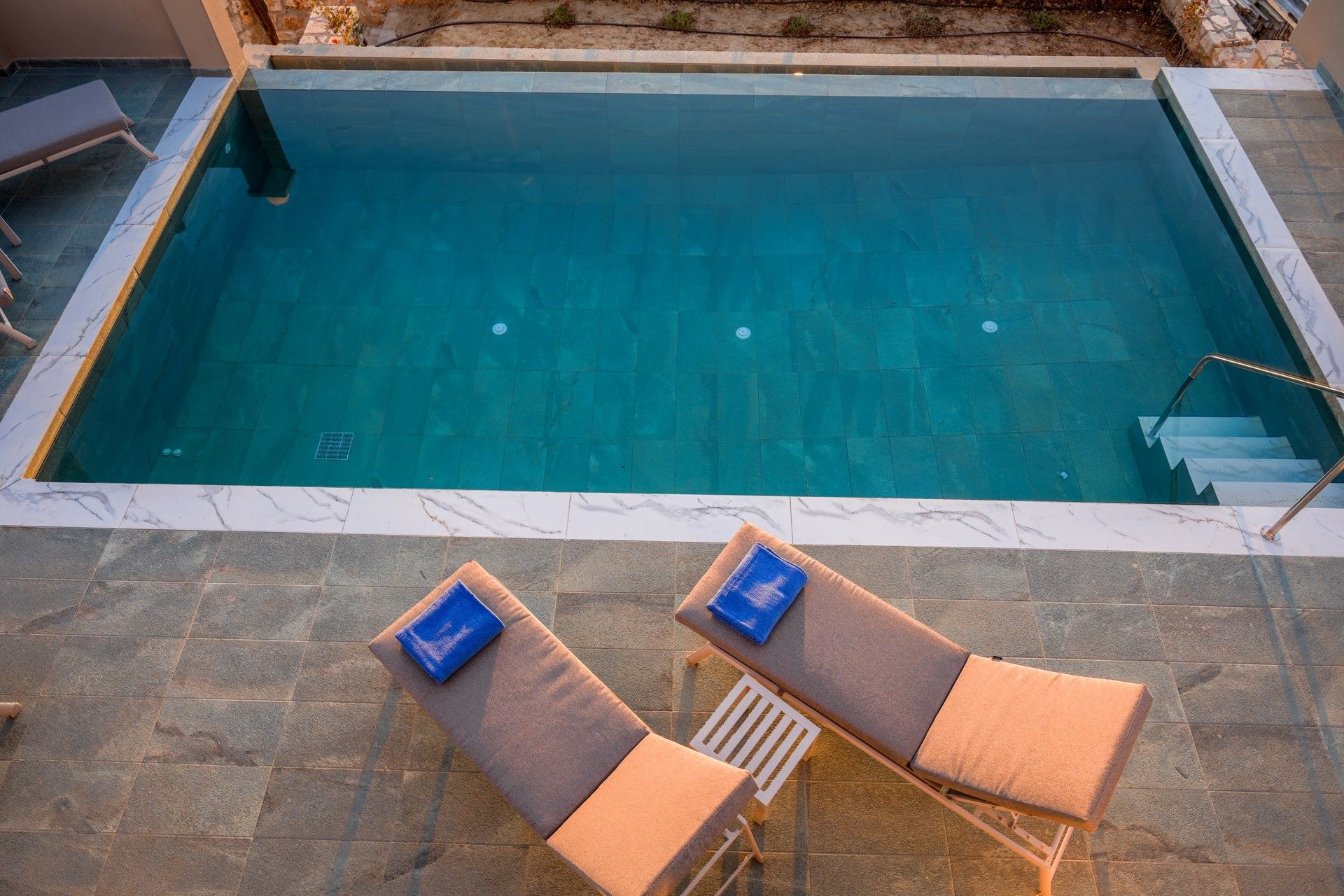 A rectangular swimming pool with a tiled edge, two lounge chairs with cushions, and a metal ladder leading into the pool.