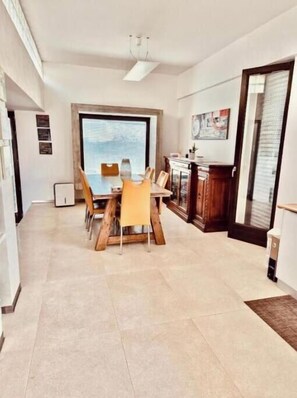 Un coin repas avec une table et des chaises, une fenêtre avec des stores, un ventilateur de plafond et un meuble sur lequel sont posés divers objets.