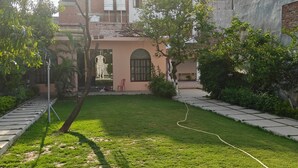 A courtyard with a lawn, a hose, and a building with a pink facade and arched windows. - Amantaran Farm Retreat (Varanasi)