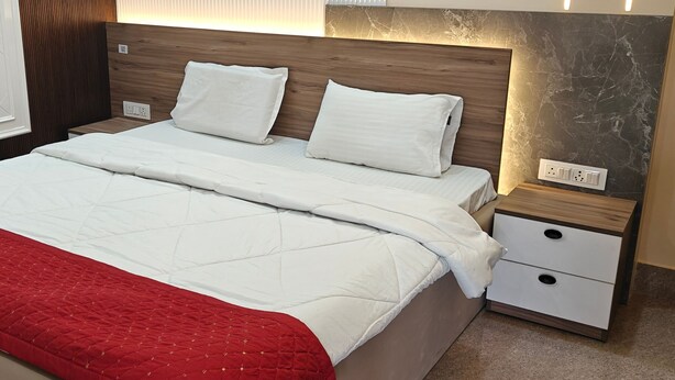 A neatly made bed with white bedding and a red blanket, a wooden headboard, a nightstand with a drawer, and a marble wall. - Amantaran Farm Retreat (Varanasi)