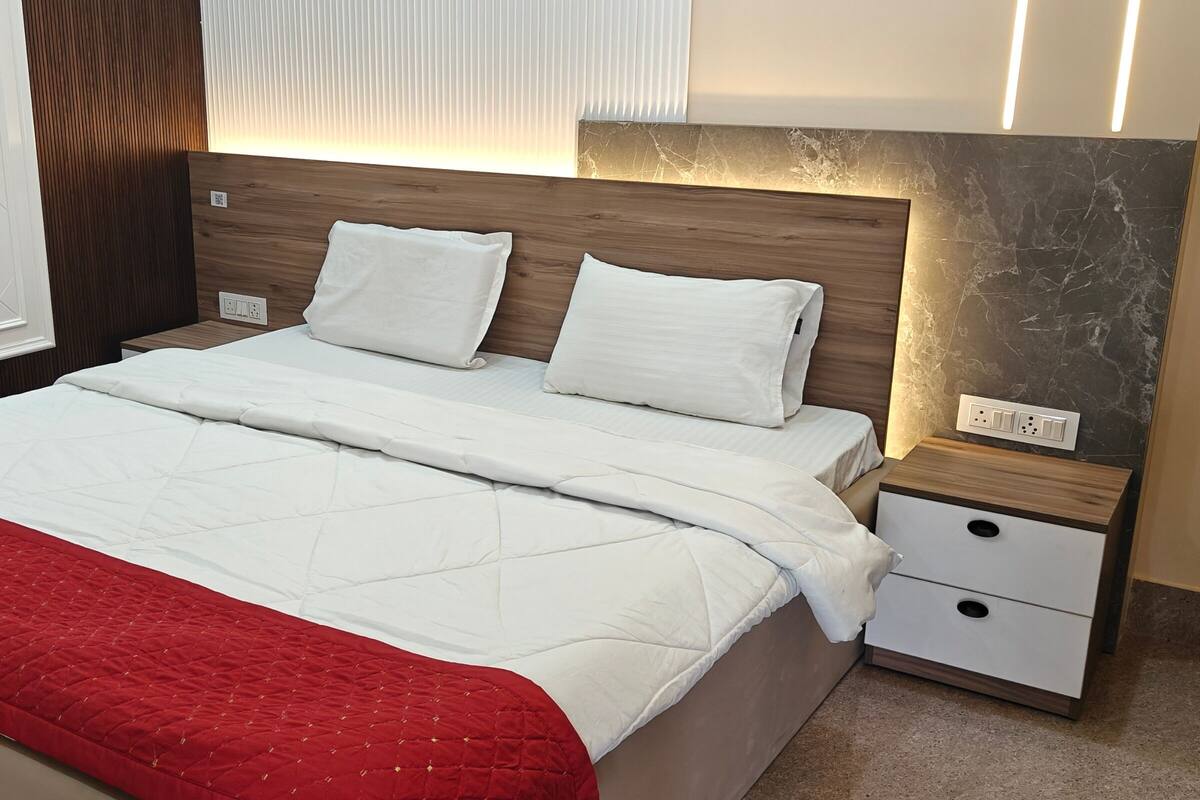 Una cama bien tendida con ropa blanca y una colcha roja, un cabecero de madera, una mesita de noche con cajón y una pared de mármol.