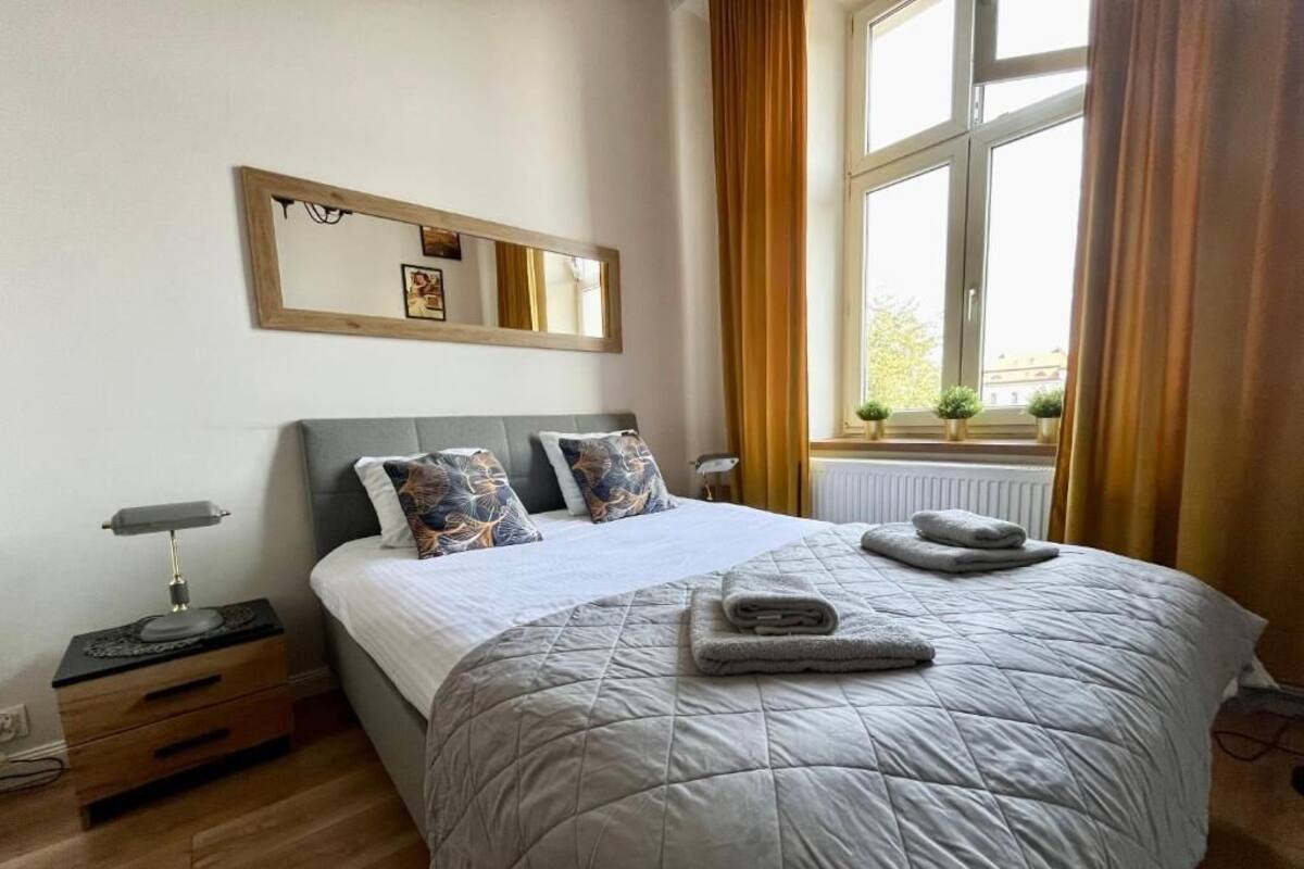 A bedroom with a bed, bedside tables, a nightstand, a window with curtains, and a wall-mounted mirror.