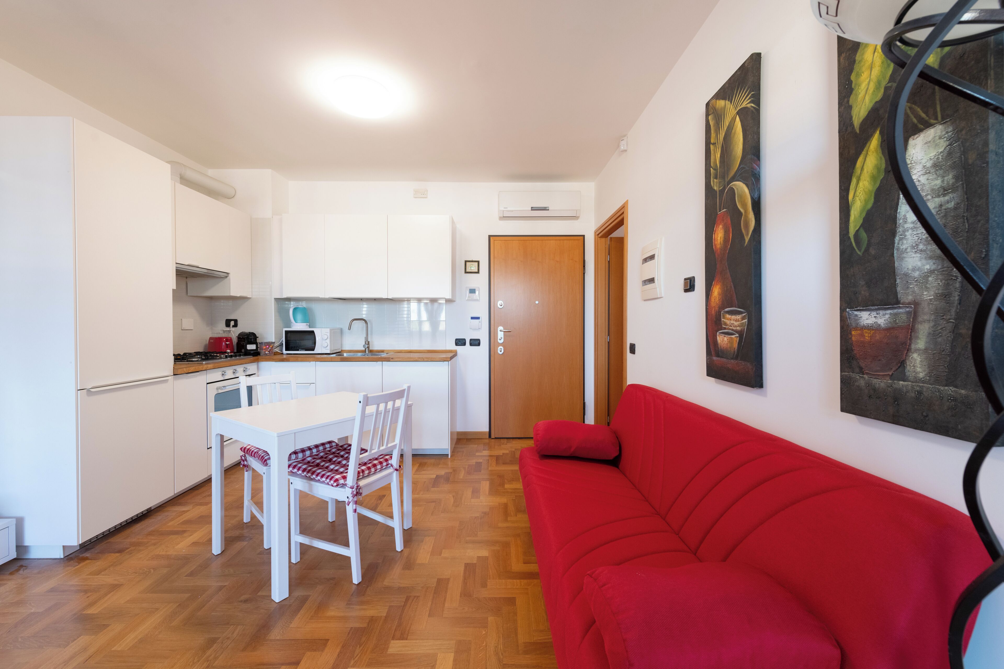 A compact kitchen with white cabinets, a dining table with chairs, and a red sofa.