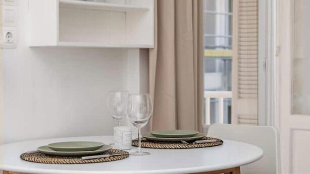 A dining table with plates, glasses, and a candle, set against a white wall with a plant on a shelf above. - Charming Couples' Apartment in Zakynthos Town (Zakinthos)