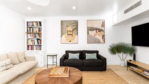 A living room with a sofa, a coffee table with a chess set, a bookshelf, and two paintings on the wall.