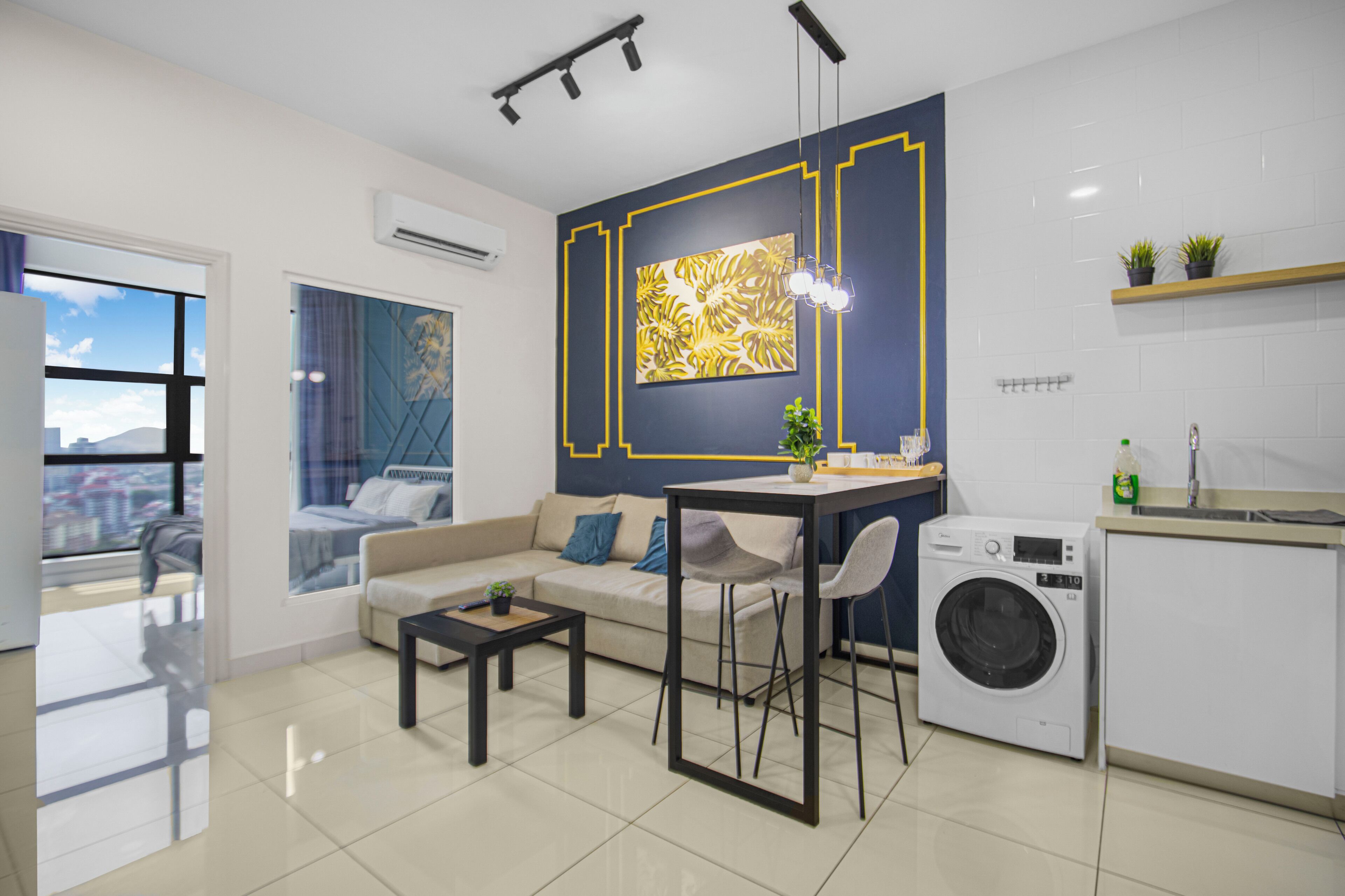 A modern living room with a sofa, coffee table, and a dining area. There is a washing machine and a small kitchen area with a sink and countertop.
