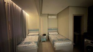 A small, compact hotel room with two single beds, a bedside table with a lamp, an air conditioning unit, and a washing machine.