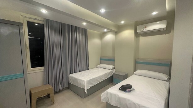 A hotel room with two single beds, a small bench, an air conditioning unit, and a window with curtains.