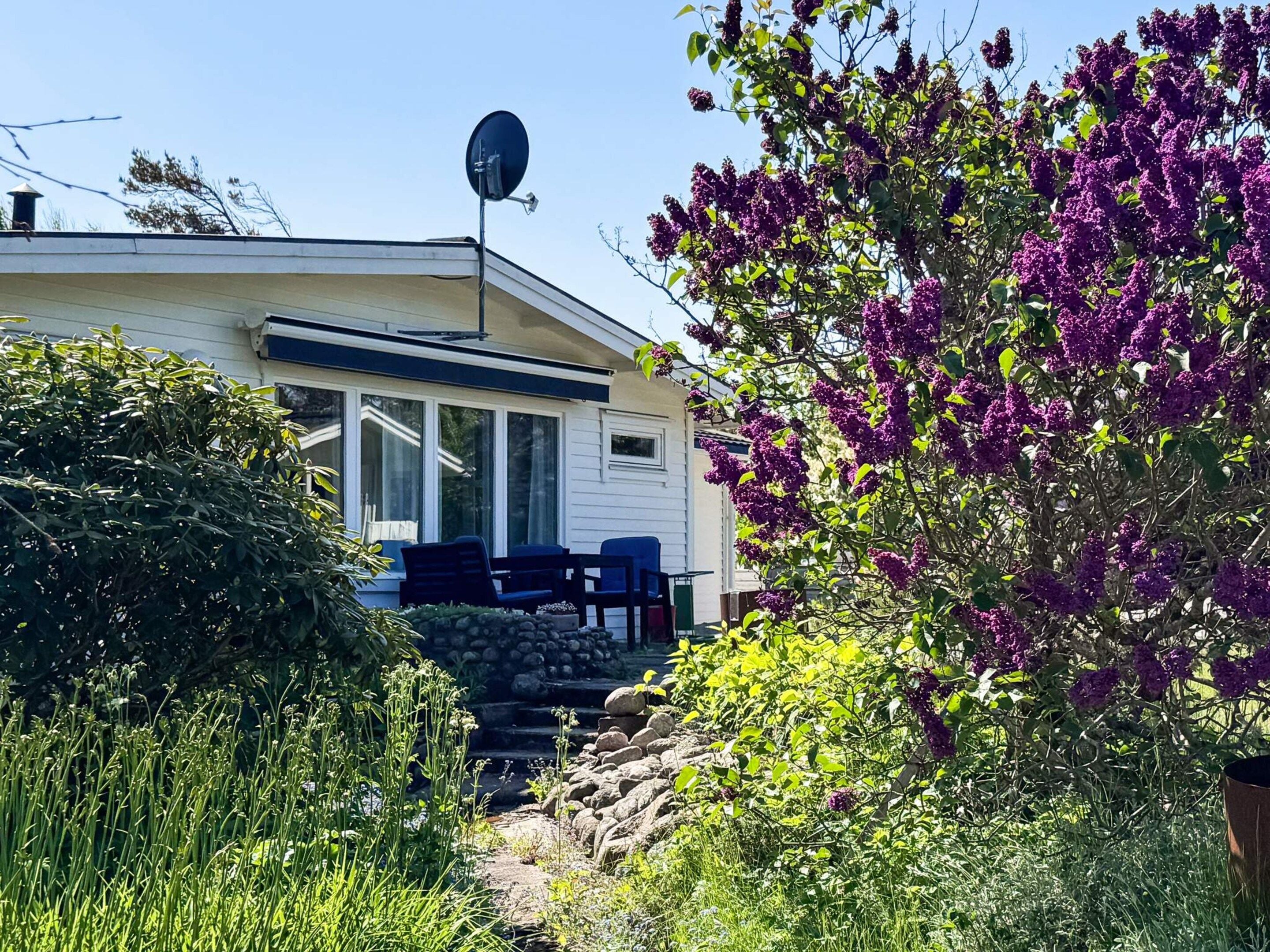 Une maison mobile équipée d’une antenne parabolique, d’une terrasse avec des chaises et d’un jardin agrémenté de fleurs violettes.