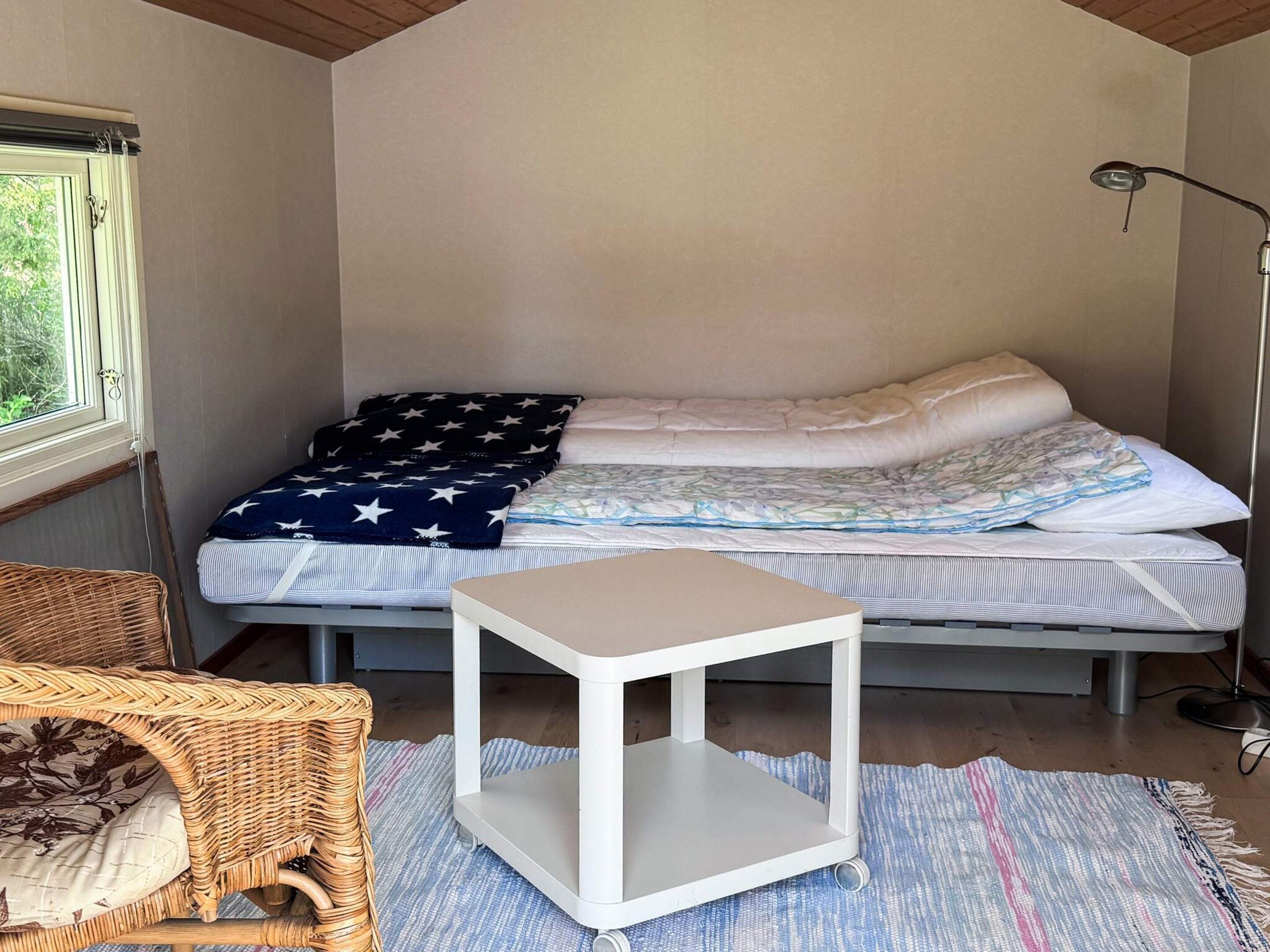 Ein kleiner, gemütlicher Raum mit einem Bett, einem kleinen weißen Tisch, einem Korbstuhl und einer Stehlampe.