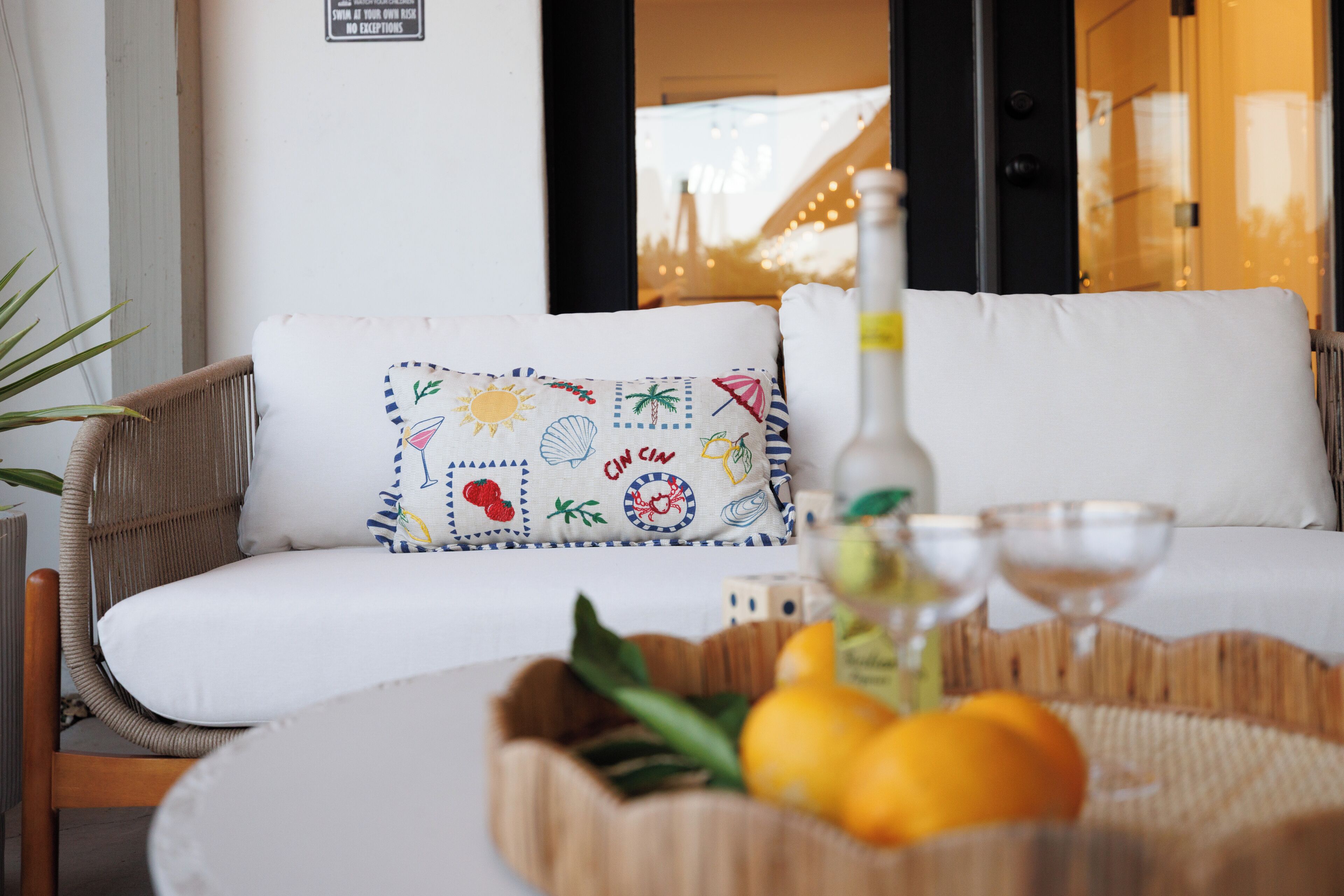 Una silla de mimbre con un cojín blanco, una cesta de mimbre con limones y una botella, y una pared con un cartel.