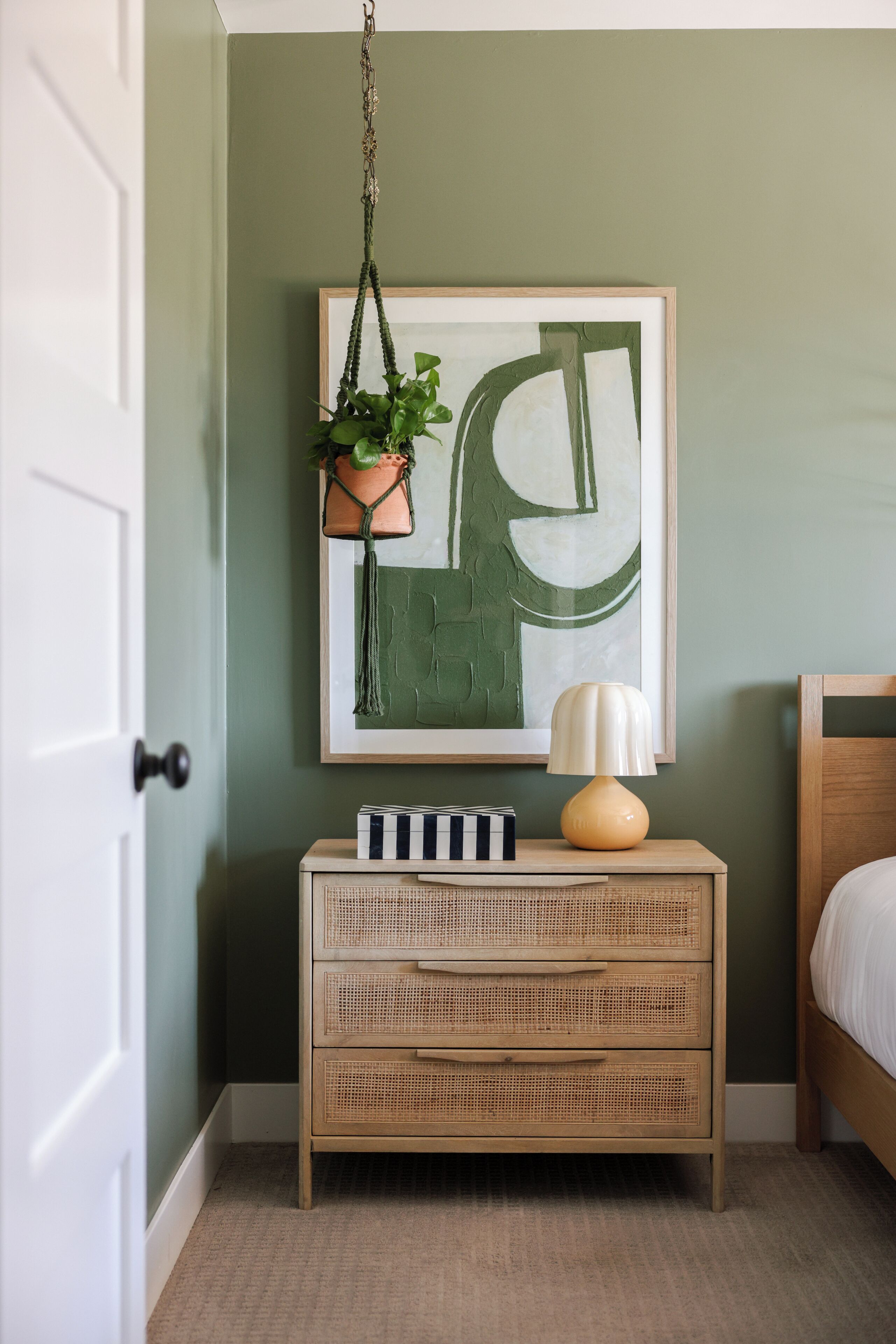 A room with a green wall, a framed abstract painting, a wicker nightstand with drawers, a hanging plant, and a bedside lamp.