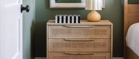 A room with a green wall, a framed abstract painting, a wicker nightstand with drawers, a hanging plant, and a bedside lamp.