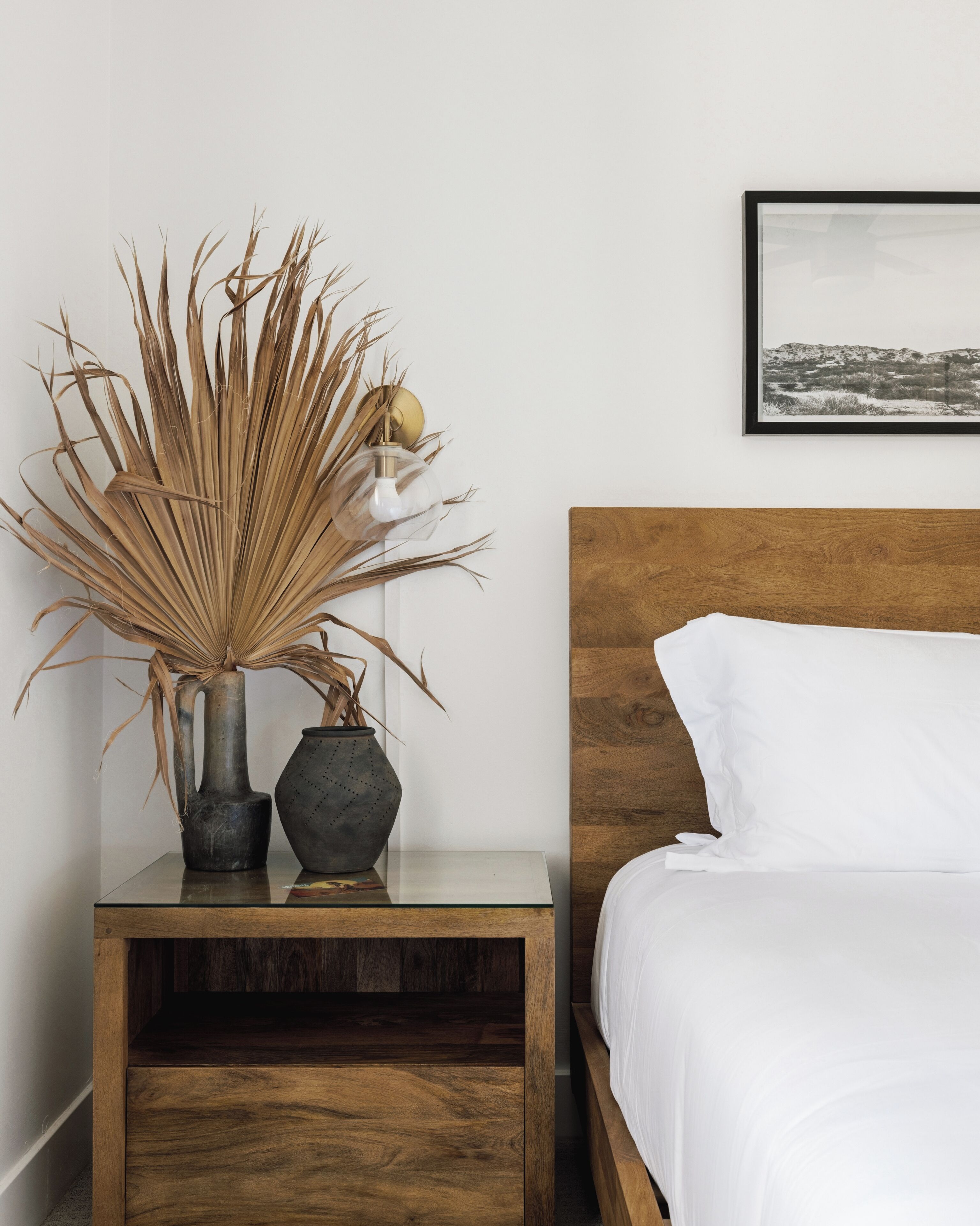 A neatly arranged bedroom with a wooden headboard, a bedside table with two vases, and a framed picture on the wall.