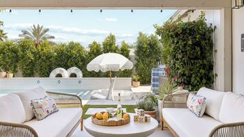 An outdoor seating area with wicker furniture, a round table with fruits and a bottle, and a white umbrella.