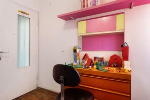 Une pièce avec un bureau en bois, une chaise, une fenêtre avec des stores et des constructions colorées en LEGO accrochées au mur.