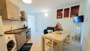 Een praktische keuken met een wasmachine, een eethoek met een tafel en stoelen, en een aan de muur bevestigde televisie.