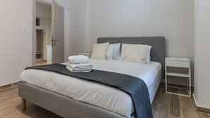 Een net opgemaakt bed met wit en grijs beddengoed, een nachtkastje en een deur naar een andere kamer.