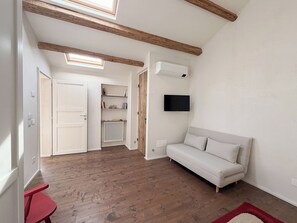 Ein Zimmer mit Holzboden, einem weißen Sofa, einem roten Tisch, einem an der Wand befestigten Fernseher und einem Oberlicht.