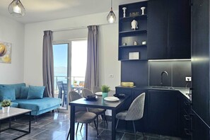 A modern living space with a sofa, dining table, chairs, a kitchen area, and a view through a large window. - Christine Sunny Corner 004 /w Sea View Balcony (Sarandë)