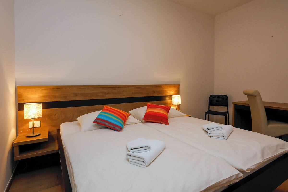 A hotel room with a double bed, two bedside lamps, a wooden headboard, a chair, and a small table.