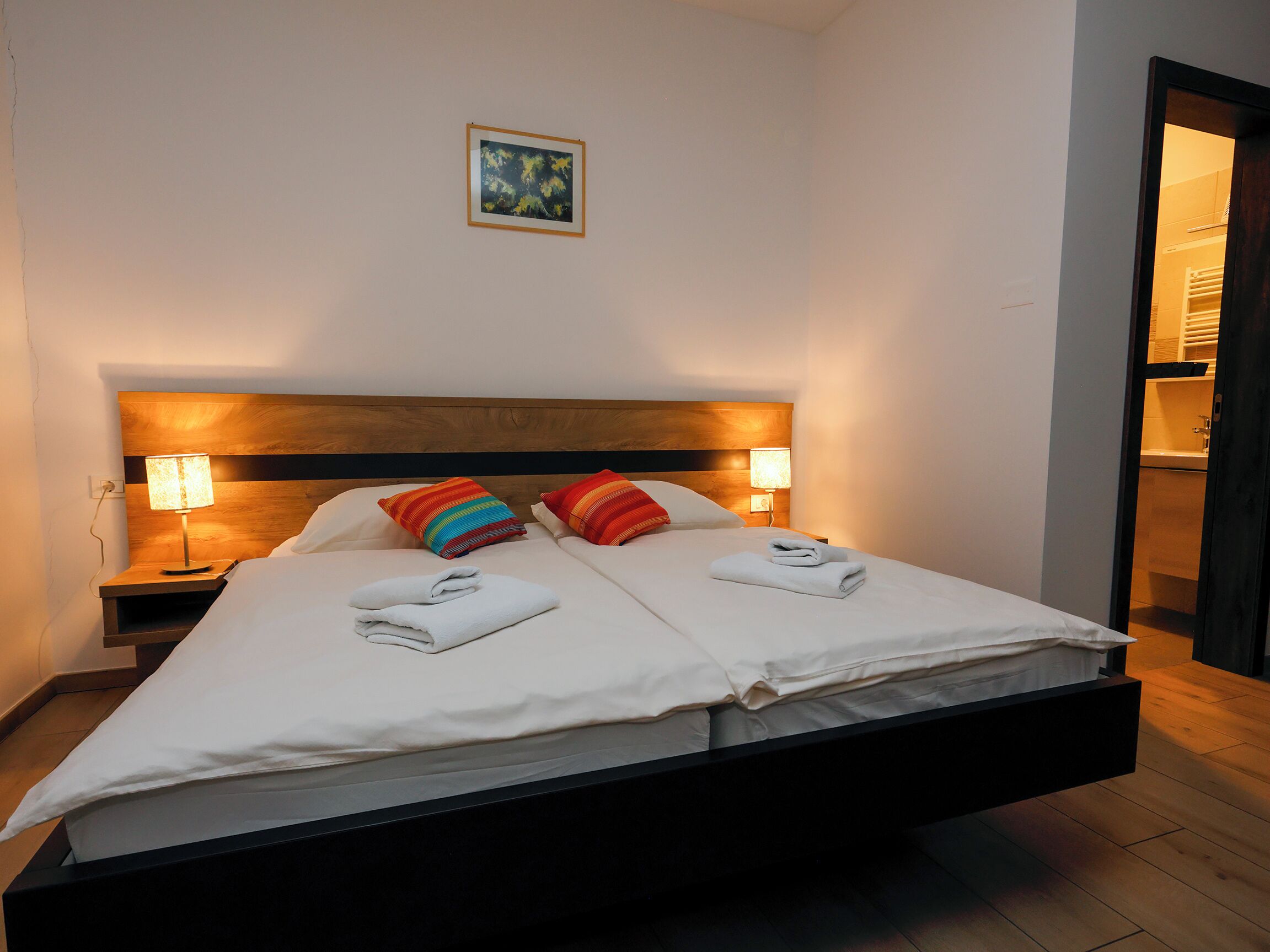 Ein modernes Hotelzimmer mit einem Bett, zwei Nachttischlampen, einem Holzkopfteil und einem kleinen Gemälde an der Wand.
