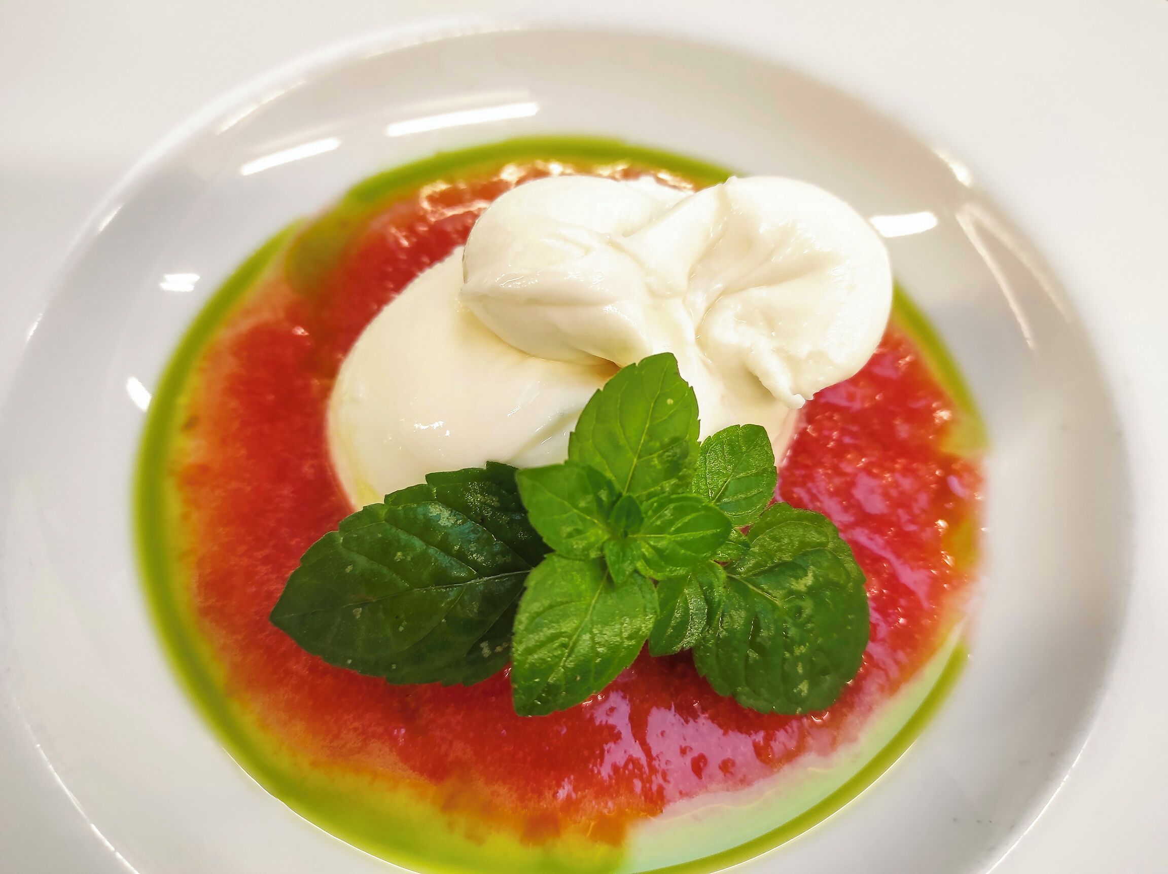 A white plate with a dessert consisting of a dollop of cream, a slice of red fruit, and green leaves.