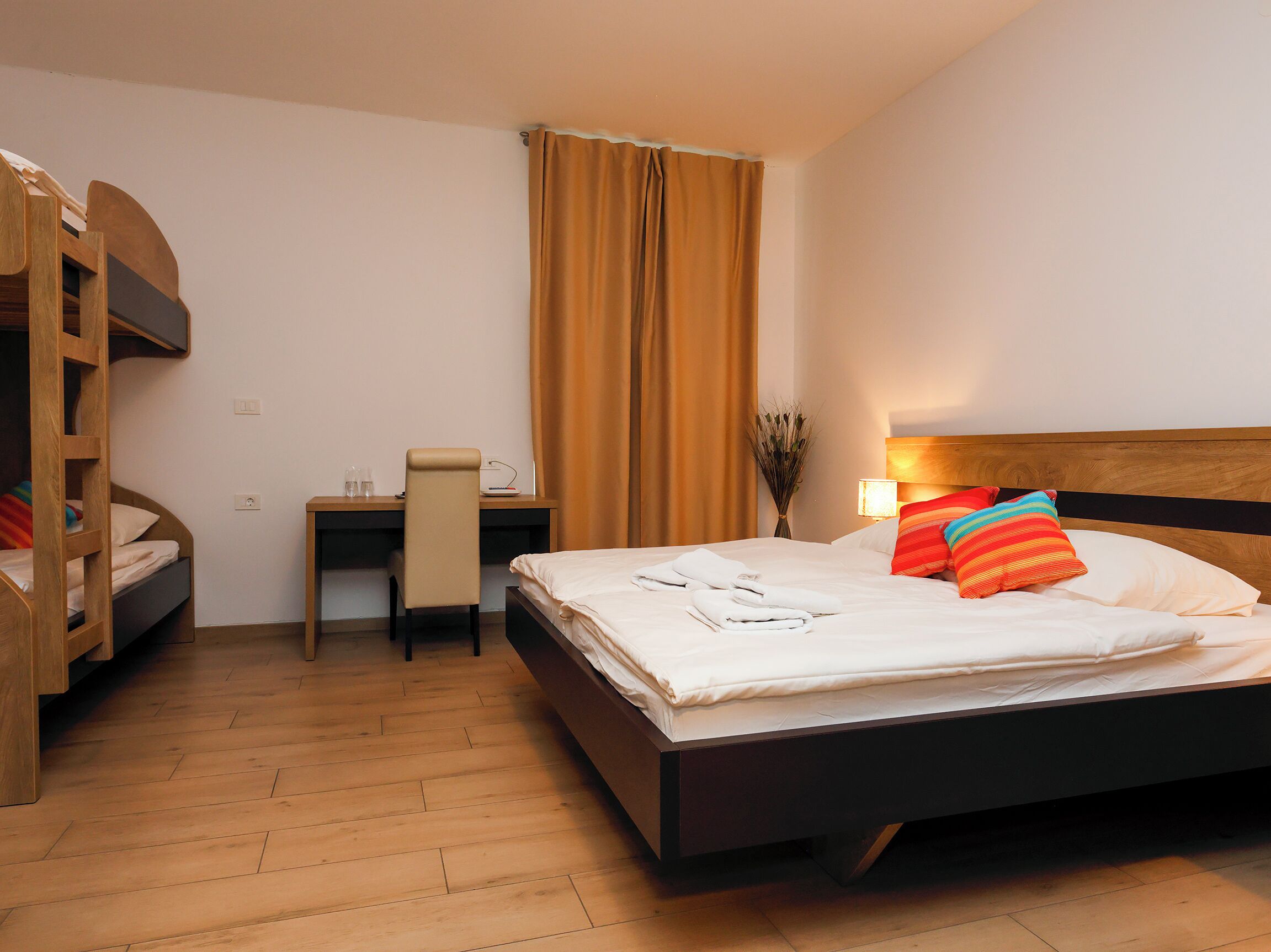 Ein Hotelzimmer mit Etagenbett, Schreibtisch mit Stuhl, Nachttisch, Lampe und einem Fenster mit Vorhängen.