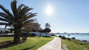 Un chemin pavé mène à un bâtiment agrémenté d’un palmier et offrant une vue sur la mer, où l’on aperçoit des bateaux.