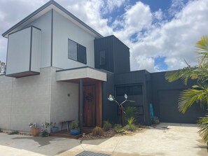 Ein modernes, zweistöckiges Haus mit einer Betonfassade, einer Holztür und einem kleinen Gartenbereich mit Pflanzen und einem dekorativen Metallstück.