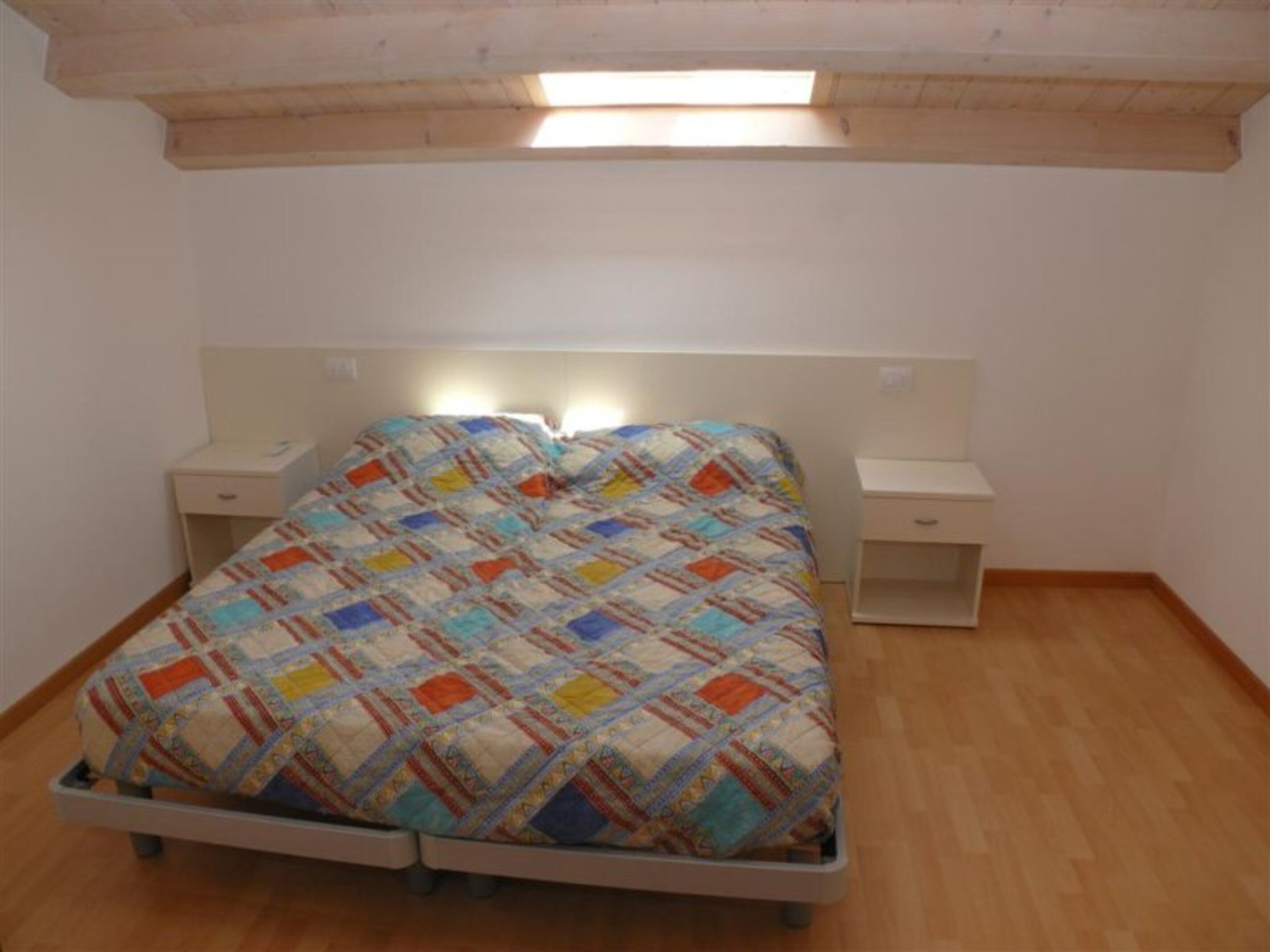Een eenpersoonsbed met een kleurrijke quilt, twee nachtkastjes en een dakvenster erboven.