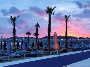 Un paseo marítimo en la playa con tumbonas, sombrillas y palmeras al atardecer.