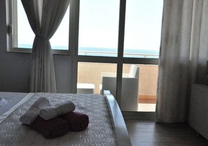 Habitación de hotel con cama, ventana con cortinas, vista al mar y balcón con una silla.