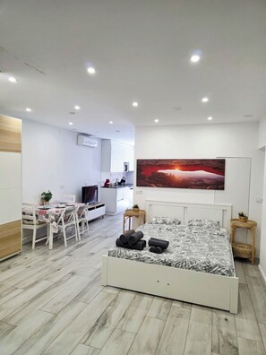 Een moderne slaapkamer met een bed, nachtkastjes, een zithoek met stoelen, een keuken achterin, en een groot schilderij aan de muur.